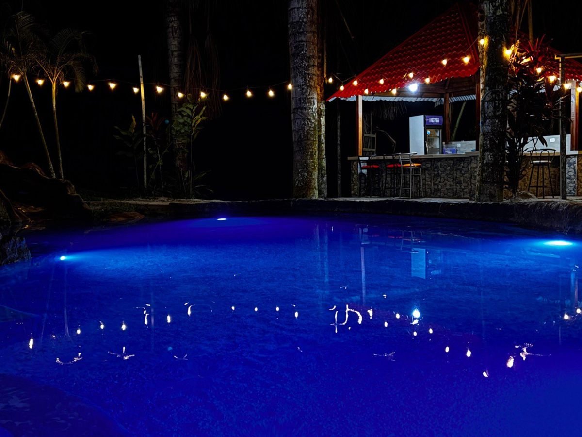 La piscina iluminada de azul por la noche, junto al bar con luces colgantes